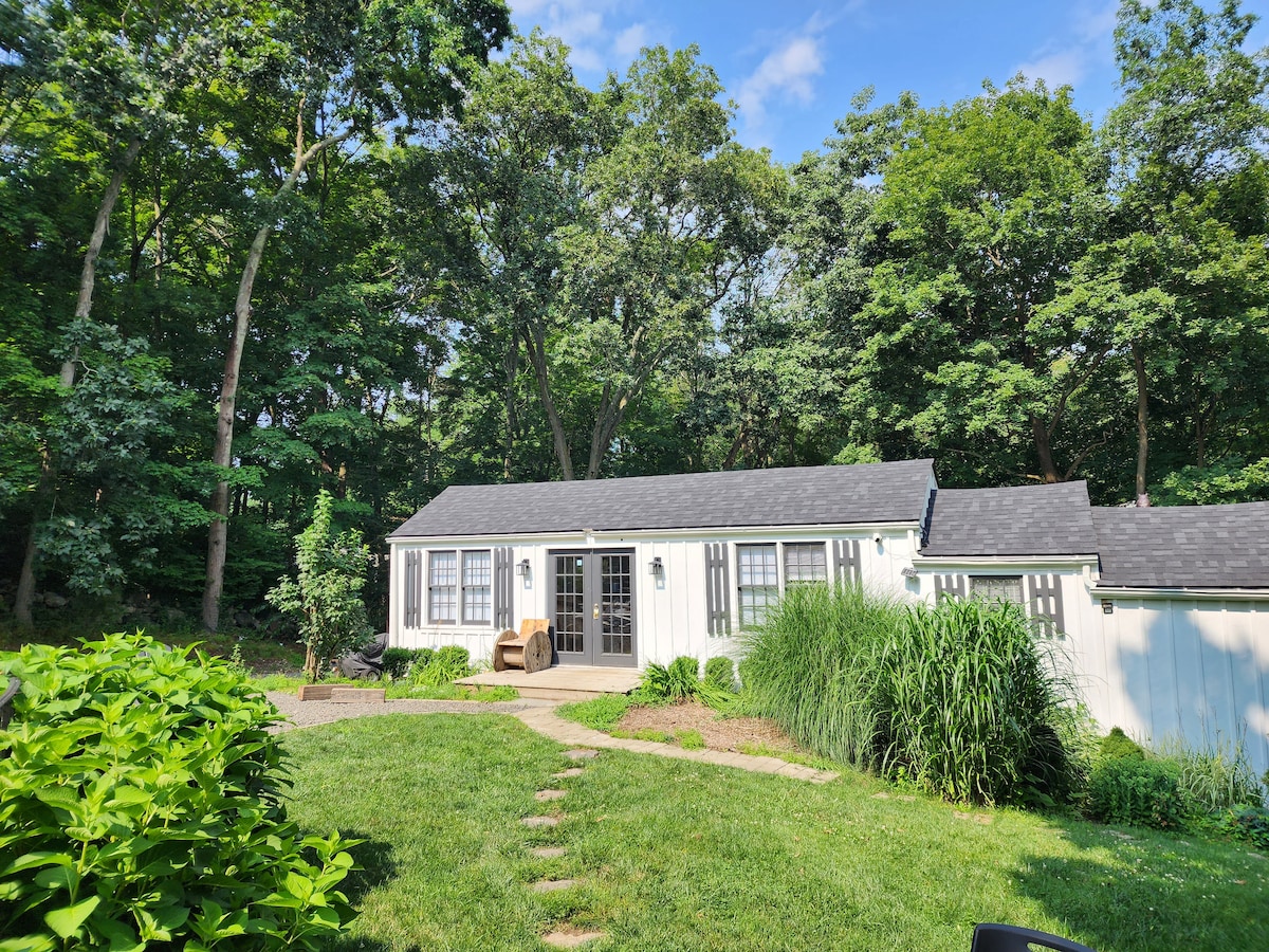 A charming single-story house is surrounded by lush greenery and tall trees, providing a peaceful ambiance. The entrance features double French doors, leading to a welcoming patio area adorned with natural grass and plants, enhancing the home's connection to the serene outdoor environment.