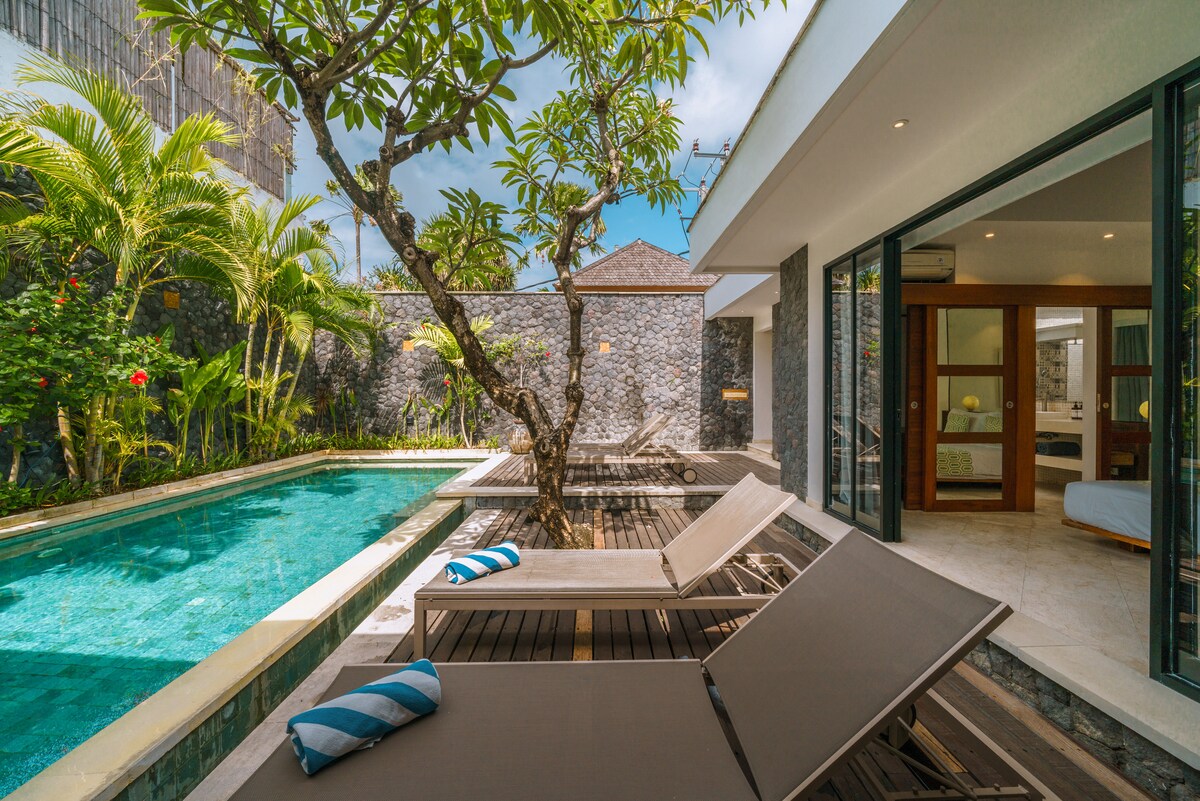 A private swimming pool is framed by lush tropical greenery, with sun loungers positioned nearby. The villa features large glass doors that create an inviting connection to the interior spaces, which are bathed in natural light.