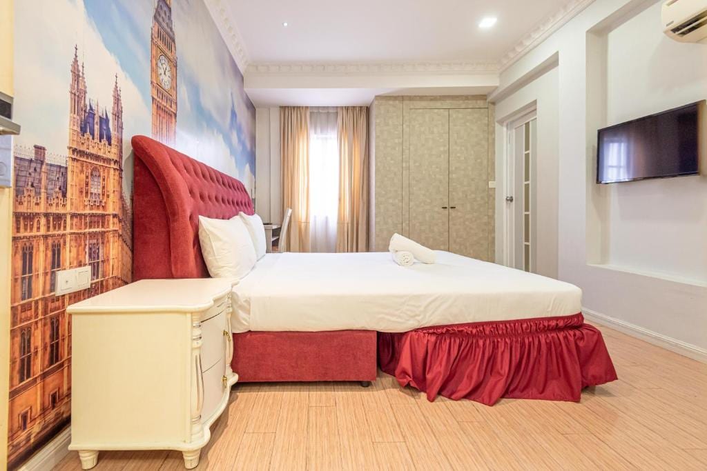 A spacious sleeping area features a large, upholstered headboard with a rich red velvet texture. The bed is covered in crisp white linens, complemented by a decorative white towel. A muted wall mural depicting a historic clock tower and light curtains allow natural light to fill the room.