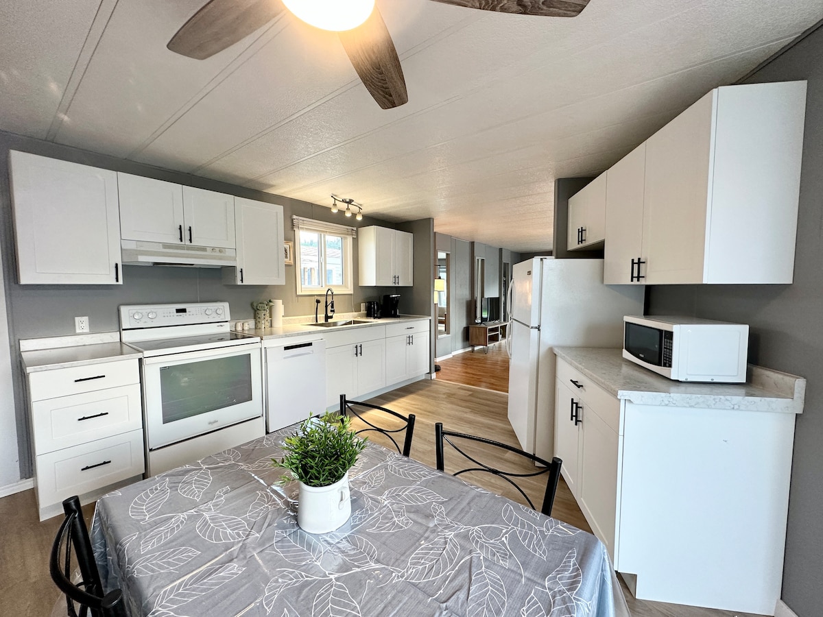 The kitchen is presented with modern white cabinetry and a full suite of appliances, including a microwave and refrigerator. A dining table with black metal chairs is positioned near a window, allowing for natural light. A ceiling fan circulates air in the open space.