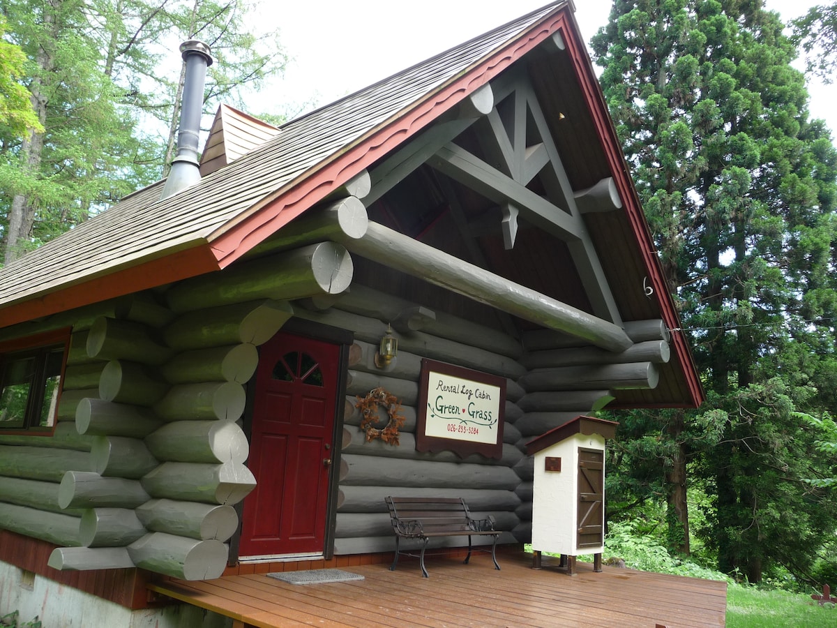 The exterior of the log cabin features a natural wood finish with a peaked roof and large beams. A red door stands out against the wooden walls, complemented by a small bench on the porch. Surrounding greenery enhances the serene setting.