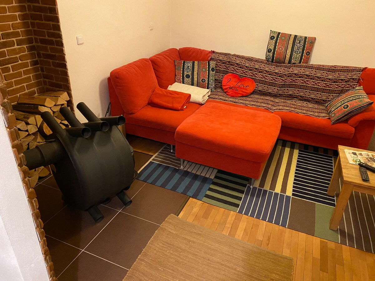 A vibrant seating area is furnished with a large red sectional sofa, complemented by a textured throw and colorful patterned cushions. A coffee table with magazines is positioned nearby, while a stack of firewood is neatly arranged in the corner, highlighting a contrast of textures.