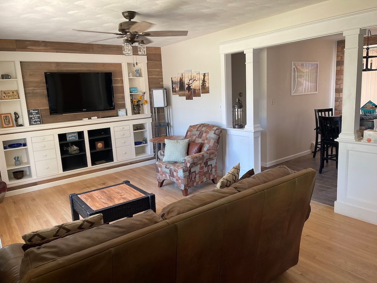 A living room area features hardwood floors and a comfortable couch, complemented by an accent chair and a wooden coffee table. Natural light fills the space, while a television is mounted on the wall. An adjacent dining area is visible through an open archway.