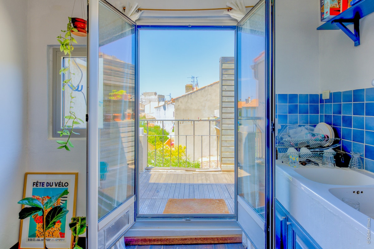 A view from an interior space shows glass doors open to a wooden deck. A blue tiled wall is visible, with a white sink and countertop on one side. Light illuminates the area, highlighting greenery outside along with city rooftops in the background.
