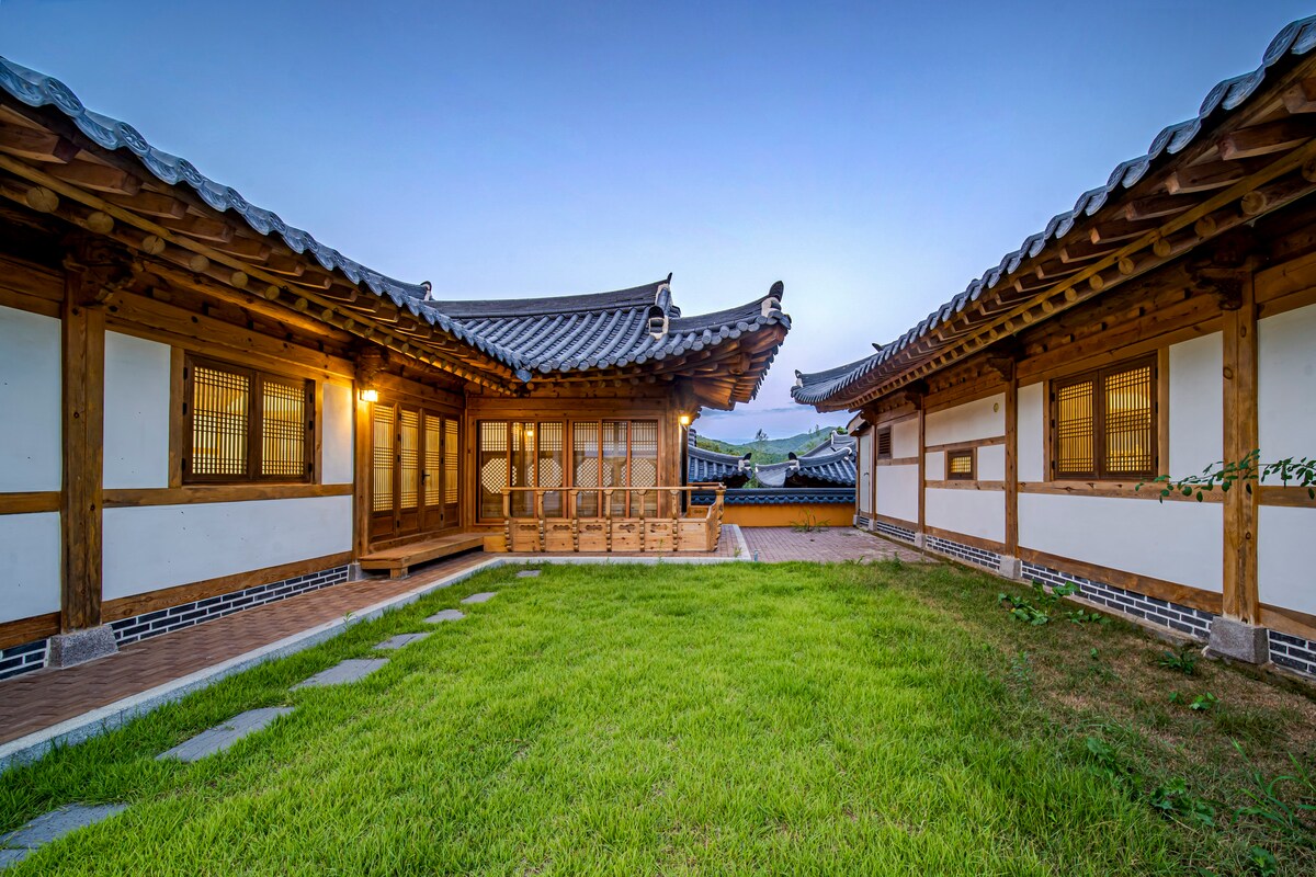 The image shows a serene courtyard surrounded by traditional Hanok buildings, featuring wooden structures with intricate eaves and large windows. Lush green grass covers the courtyard, while warm light emanates from the windows, creating a calm and inviting atmosphere in the evening light.