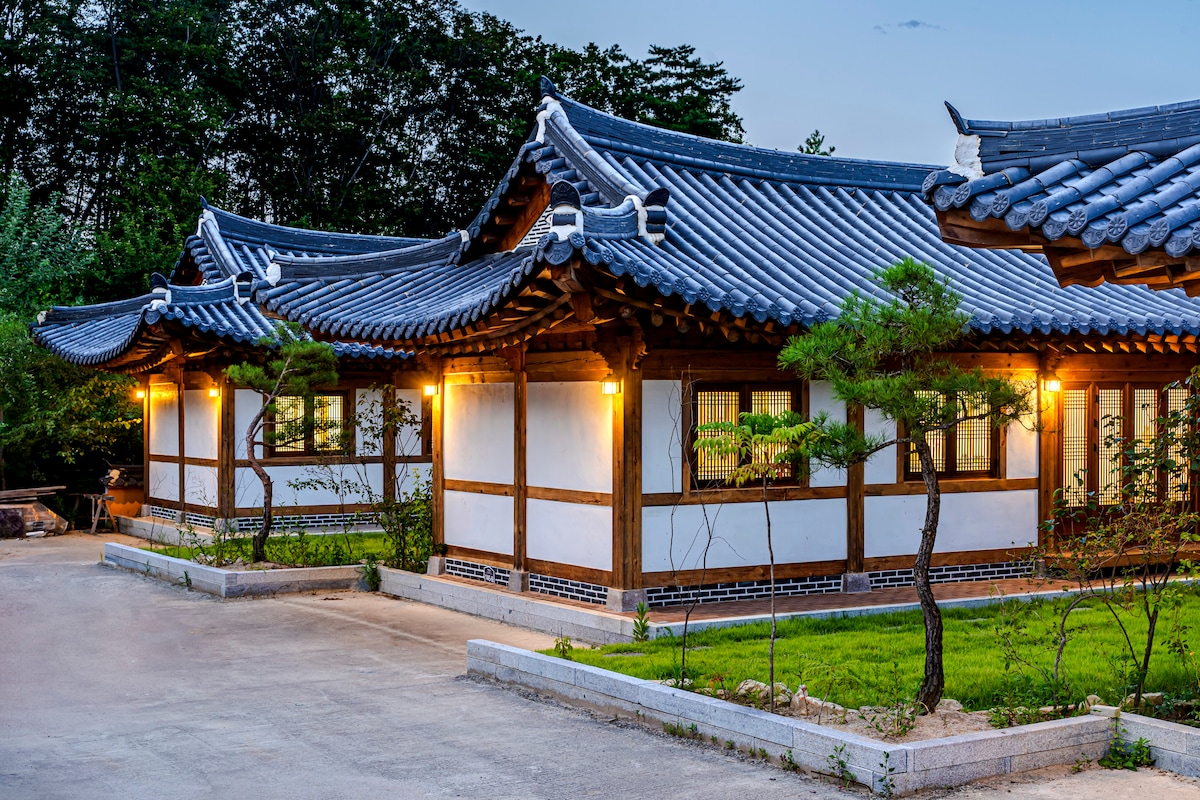 The image displays a traditional Korean hanok with gracefully curved roofs, illuminated by soft exterior lights. The structure, built with natural materials, features wooden beams and decorative windows, surrounded by manicured greenery and a well-maintained pathway.