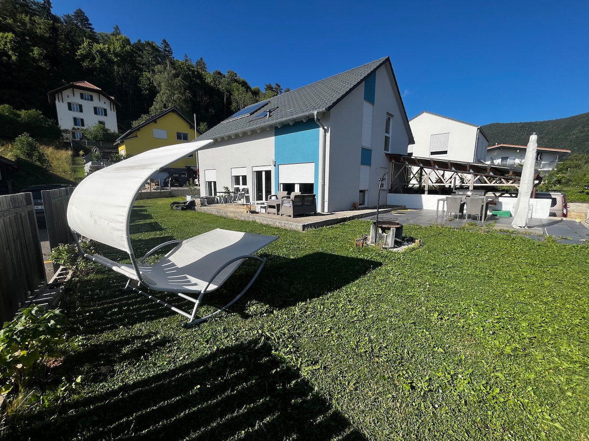 The exterior of the house is showcased, featuring a spacious grassy yard with a comfortable lounge chair and a patio area. A large deck is visible at the rear, equipped with outdoor seating. The surrounding landscape includes trees and a clear blue sky.