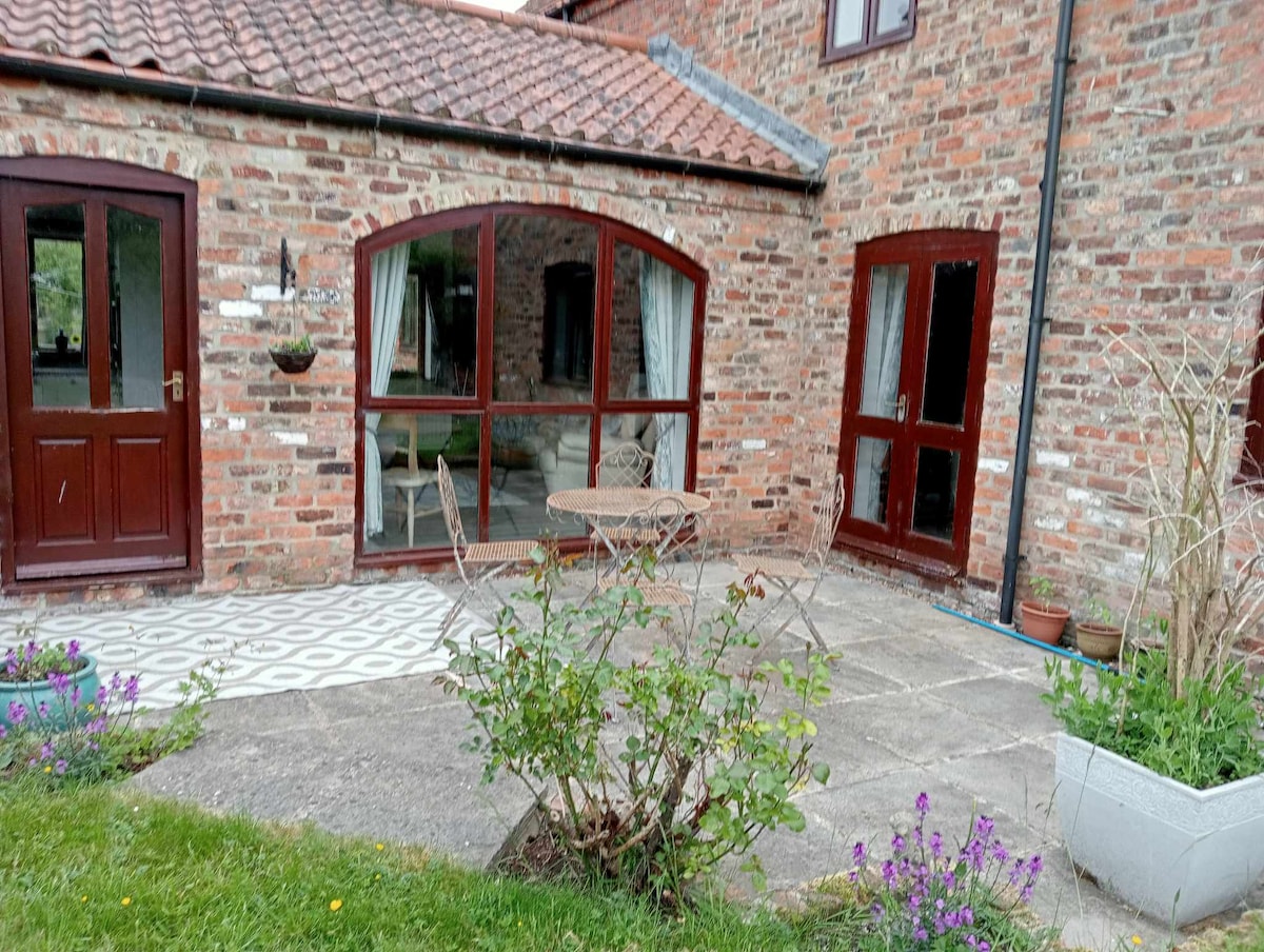 A stone patio area offers a small circular table and two chairs, surrounded by a well-maintained garden with greenery and colorful flowers. Large arched windows provide views into the interior space, showcasing a bright and inviting atmosphere while the rustic brick walls add character to the setting.