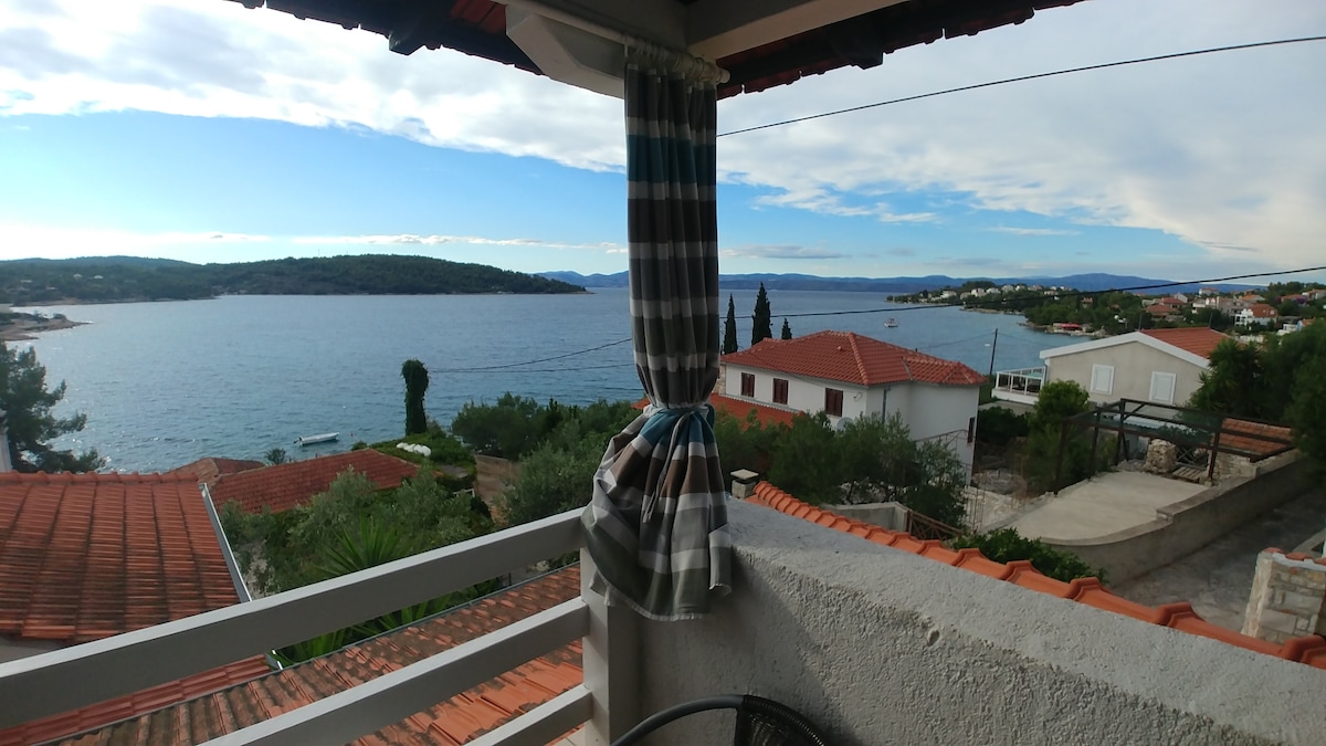 A panoramic view from the balcony captures the serene sea and lush gardens. The landscape features terracotta rooftops and green foliage, set against a clear blue sky. A patterned curtain sways gently in the breeze, framing the picturesque scene.