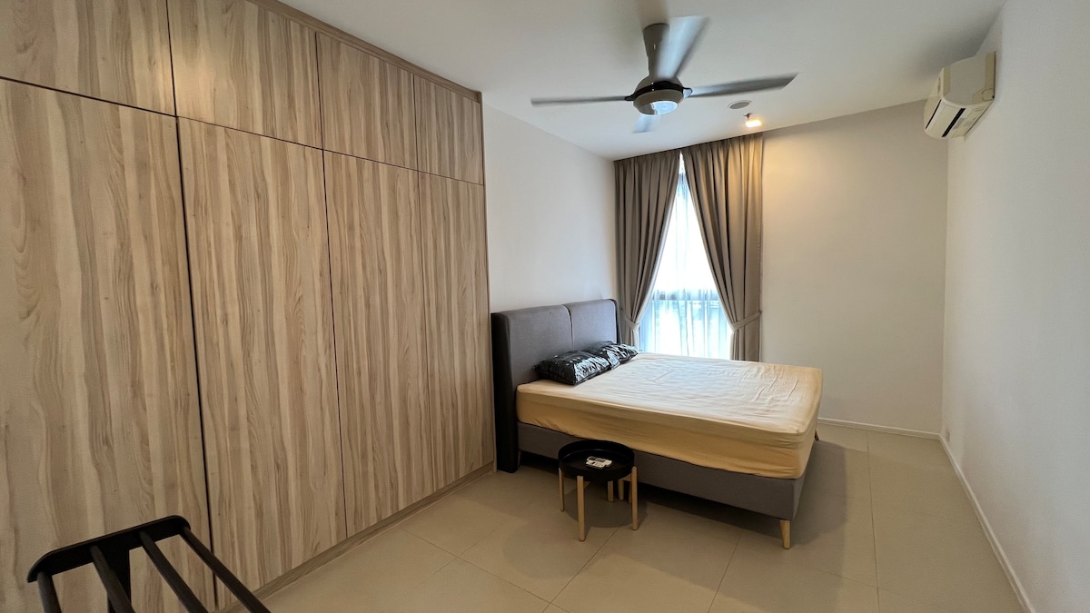 A modern bedroom is featured, showcasing a spacious bed with a light-colored mattress and two pillows. Minimalist wooden cabinetry lines one wall. Natural light filters through a large window, dressed in soft drapes. A ceiling fan is mounted for airflow, and a small stool is positioned beside the bed.
