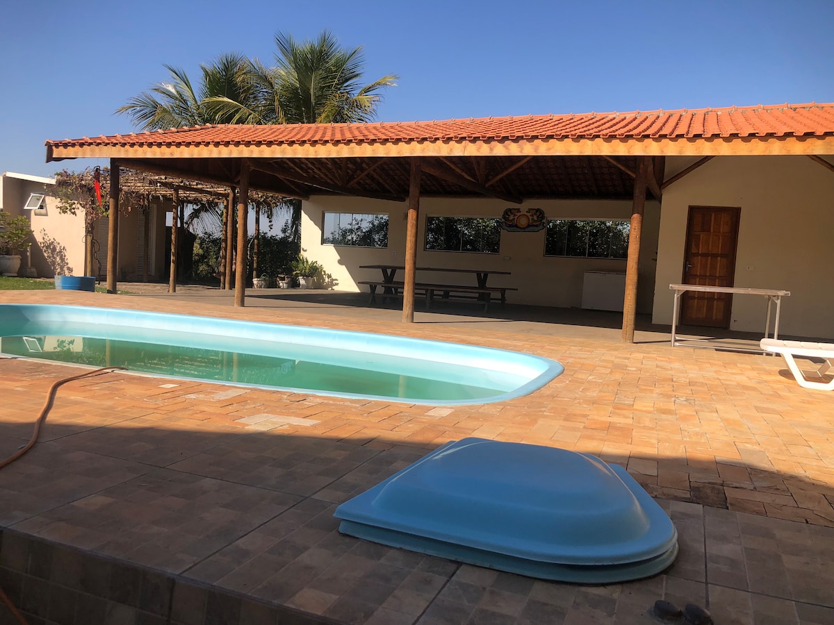 An outdoor area features a swimming pool surrounded by tiled flooring. A covered patio with wooden posts provides shade, with ample seating visible around a large table. Lush palm trees are seen in the background under clear blue skies.