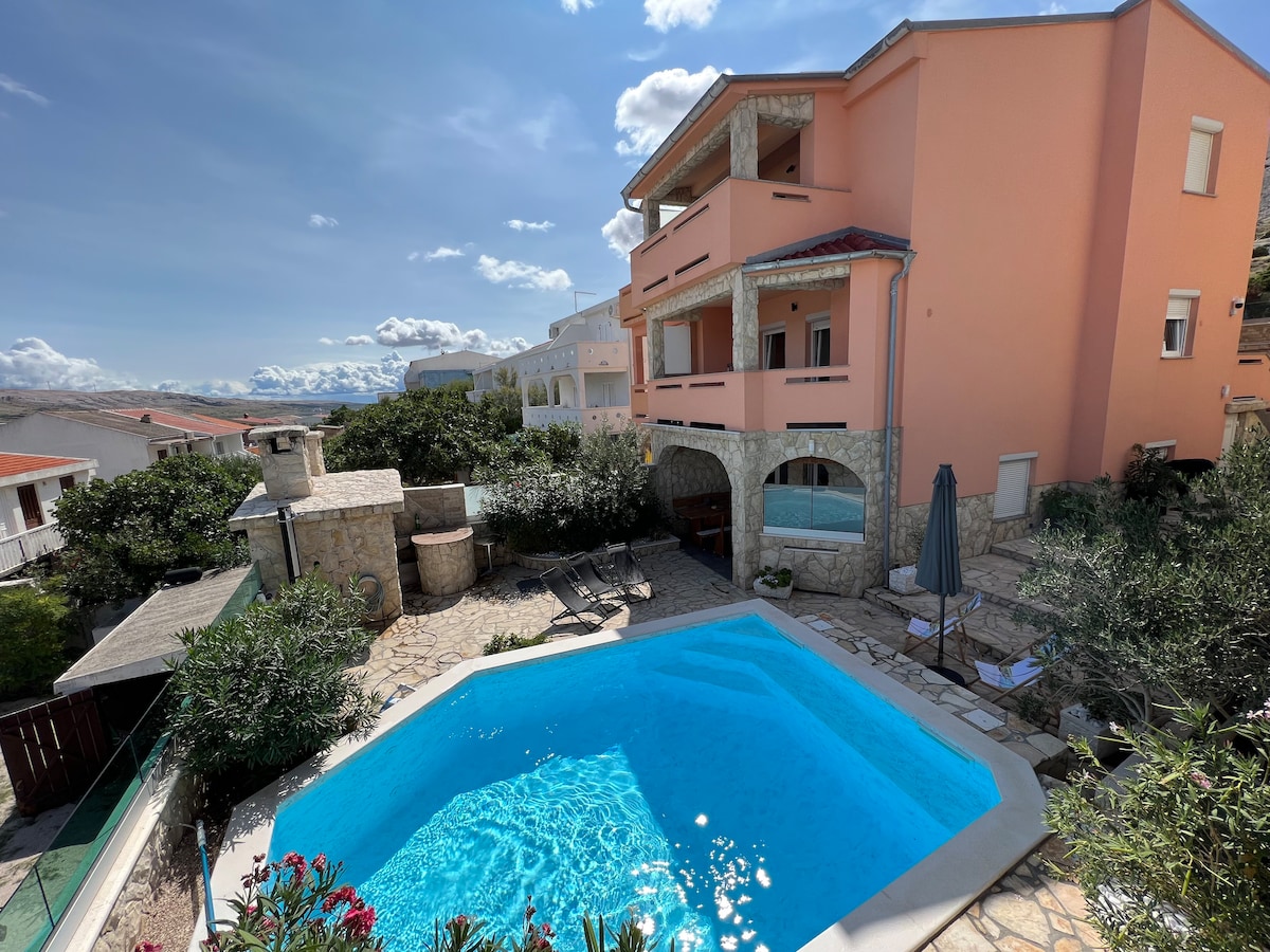 A clear blue pool is surrounded by stone paving and lush greenery. The nearby home features a light pink exterior, with multiple levels and balconies. A barbecue area and lounge chairs are positioned around the pool, inviting relaxation in an outdoor setting.