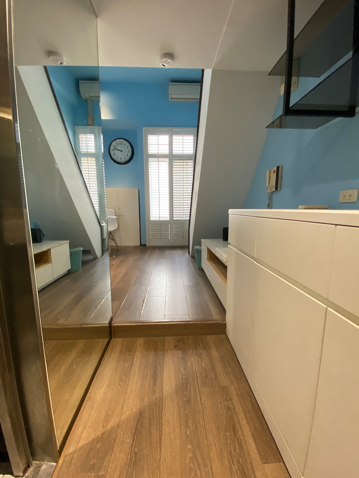 A bright entryway features a large mirror reflecting the sleek white cabinetry and warm wooden flooring. A wall clock is mounted above a small seating area, and large windows provide ample natural light, tucked beneath a staircase with a modern design.