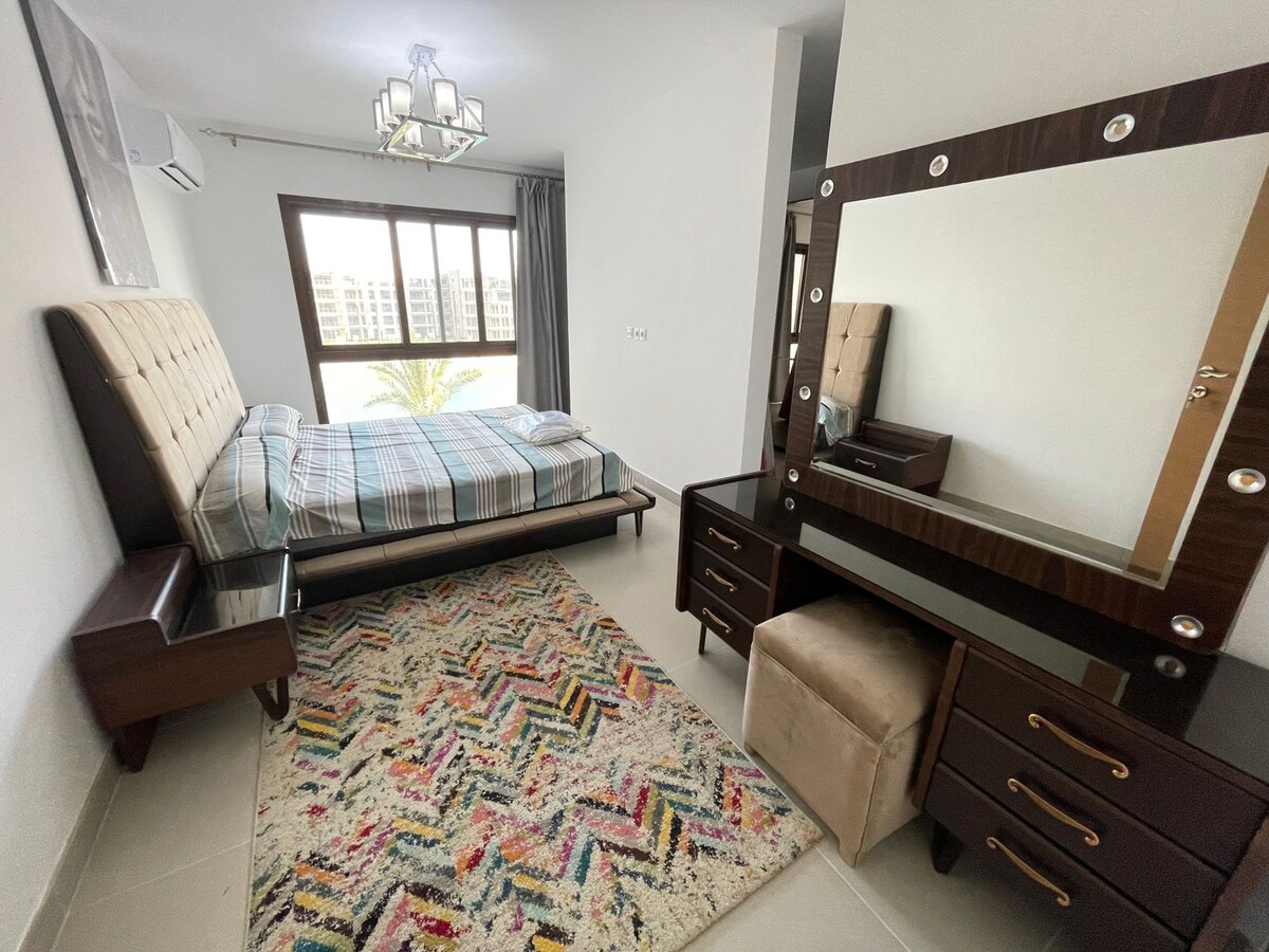 A cozy bedroom features a spacious bed with a checkered blanket and a mirrored dresser. The soft, multi-colored rug adds comfort to the area. Natural light filters through large windows, offering a view of palm trees and the surroundings.