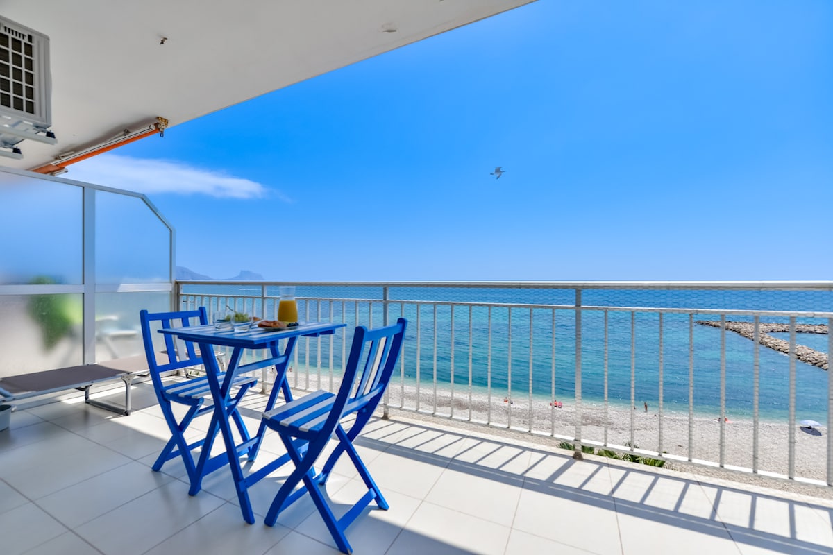 A balcony offers a serene view of the beach, where calm blue waters meet a clear sky. A blue dining set with two chairs and a table is positioned for enjoying meals outdoors. Sunlight reflects off the water, creating a peaceful ambiance.