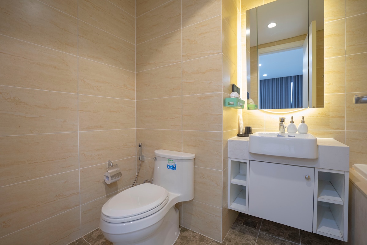 The bathroom features beige tiled walls and a light-colored vanity with a sink. A large mirror reflects the natural light, enhancing the space. Shelves above the sink hold toiletries, while a toilet is positioned beside a roll of toilet paper. The floor is covered with darker tiles.