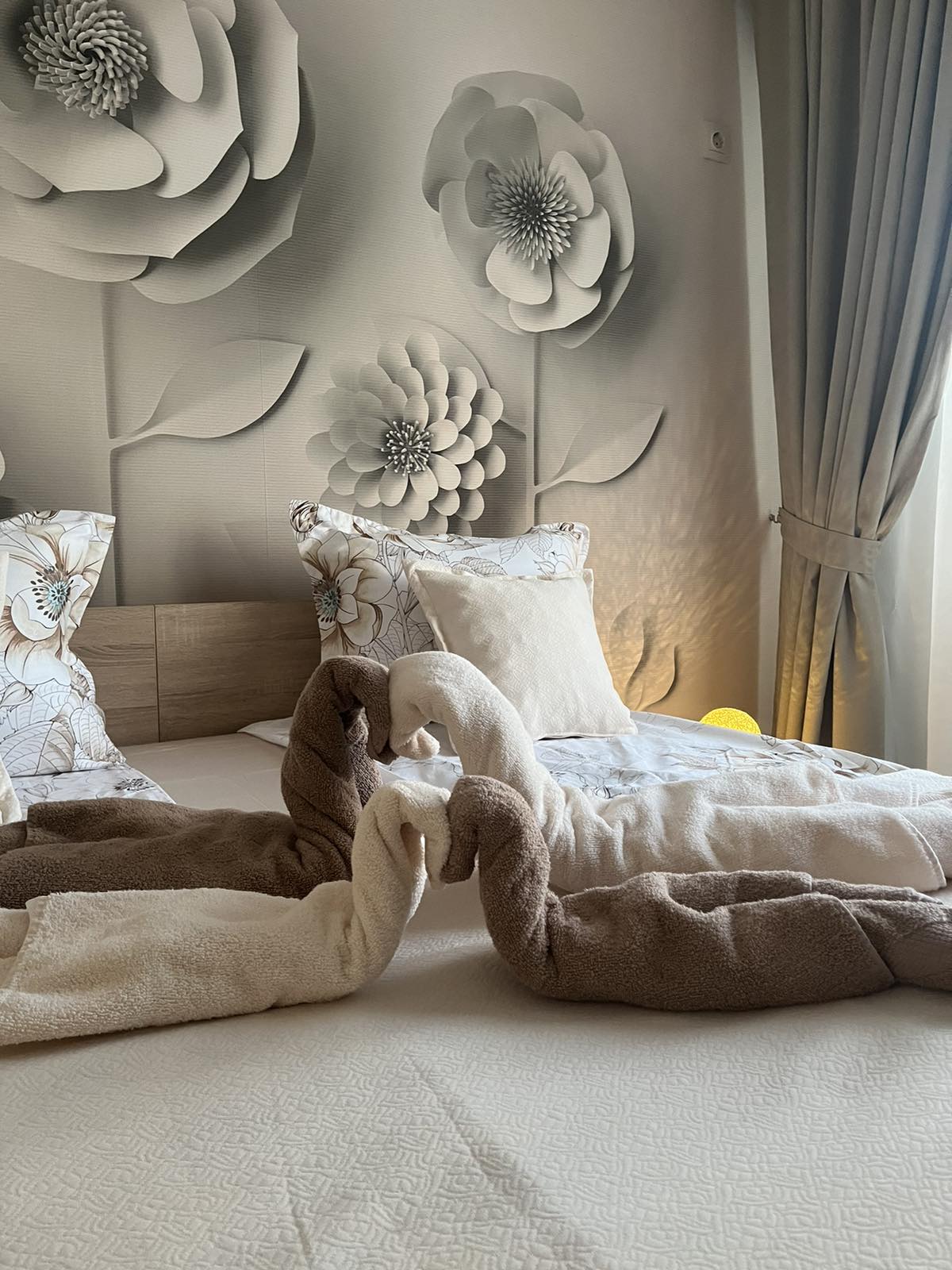 A well-appointed sleeping area is highlighted by a large floral mural on the wall. Soft pillows are placed against the headboard of the wooden bed, which is dressed in light linens. A pair of towel swans adorns the bed, creating an inviting touch.