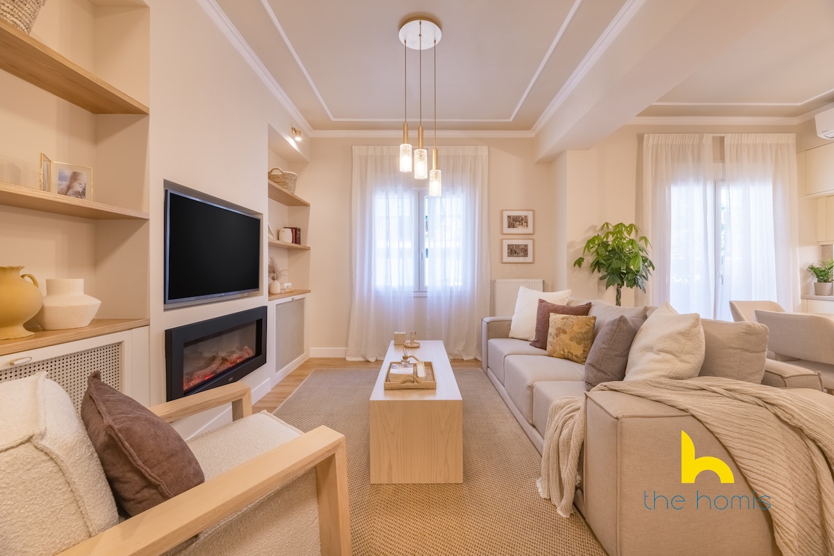 A spacious living room is furnished with comfortable seating, including a cozy sofa and accent chairs. Natural light fills the space through large windows, and an electric fireplace adds warmth. Neutral tones are complemented by soft textiles and subtle decor elements, creating a calming atmosphere.