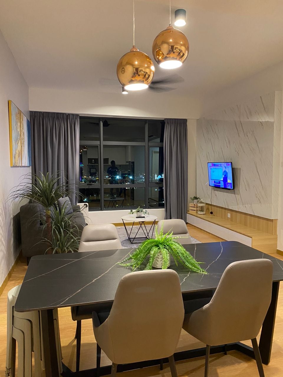 A modern living area features a sleek dining table surrounded by four light-colored chairs. Elegant metallic light fixtures hang above, while a cozy sofa and decorative plants add warmth. A large window invites natural light, showcasing a nighttime cityscape reflected in the glass.