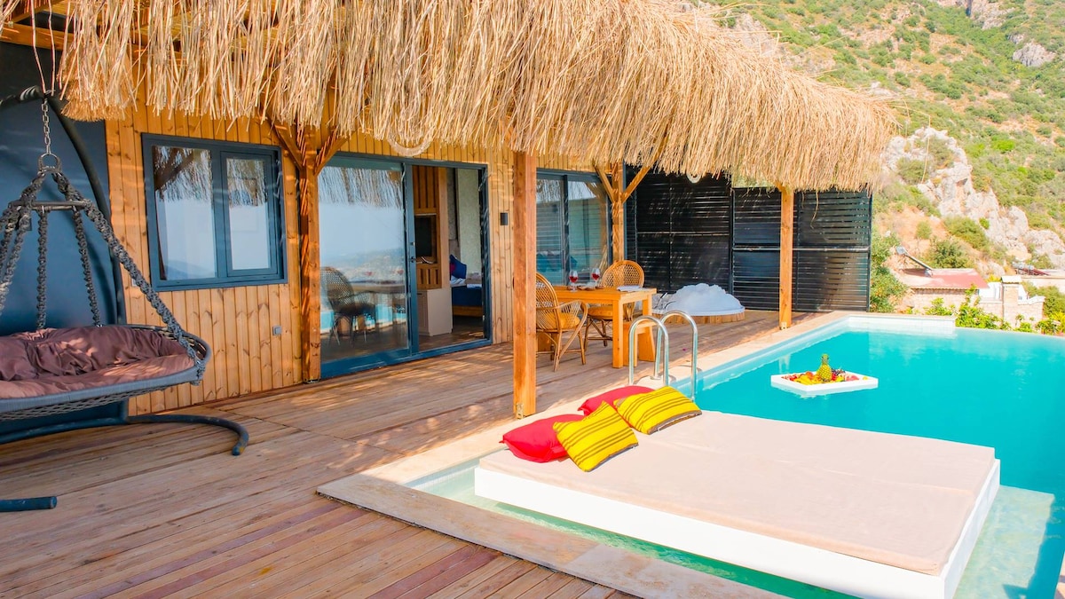 A spacious terrace features a swimming pool surrounded by wooden decking. Comfortable seating and a table for two are positioned under a traditional thatched roof. Soft cushions are placed on a sunbed, enhancing the serene outdoor space with a view of the natural surroundings.