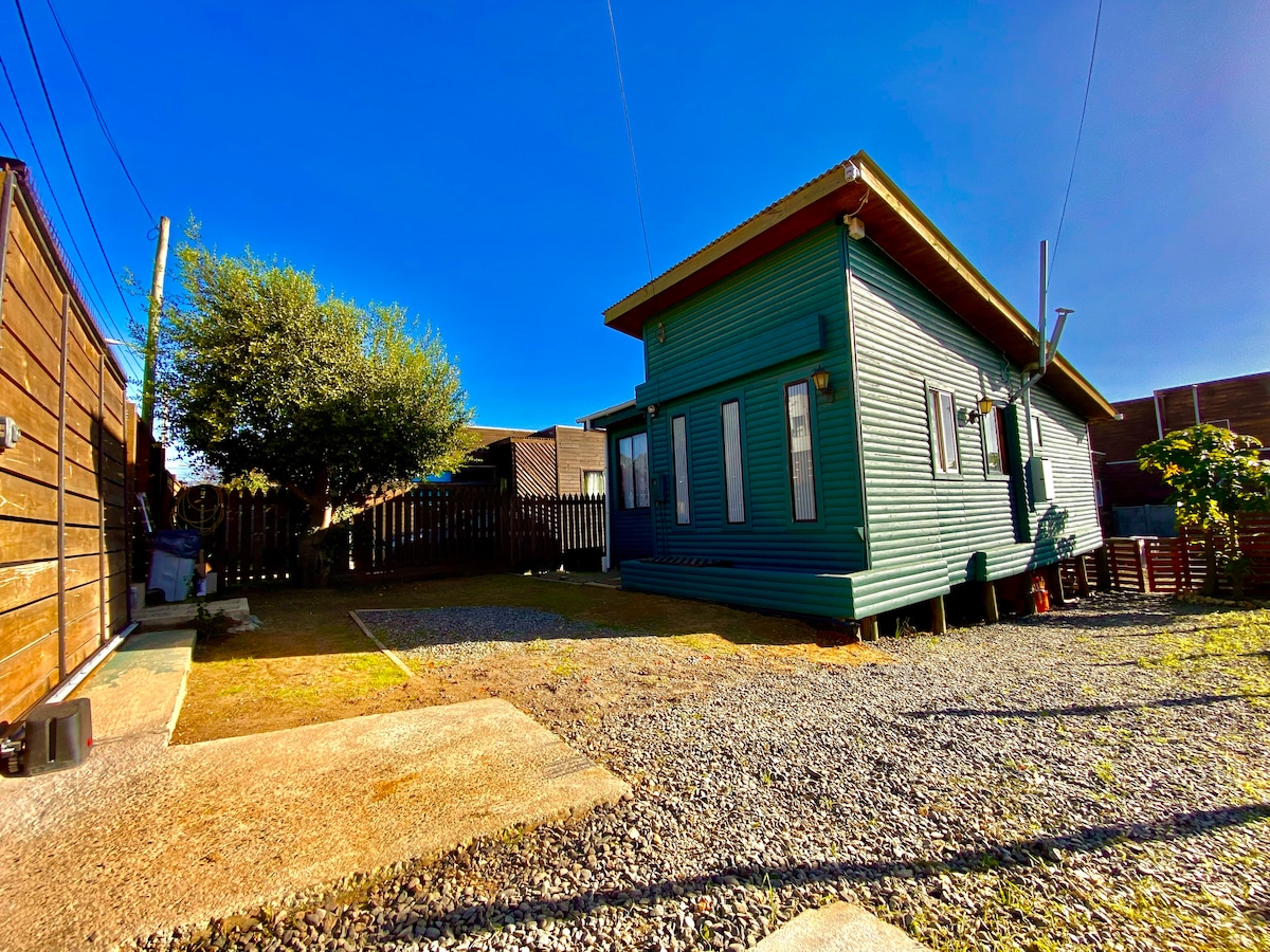 The exterior of the cabin is shown, featuring a green wood-paneled structure with multiple windows. A small yard with gravel and grass surrounds the cabin, and a tree provides shade nearby. The clear blue sky complements the outdoor setting.