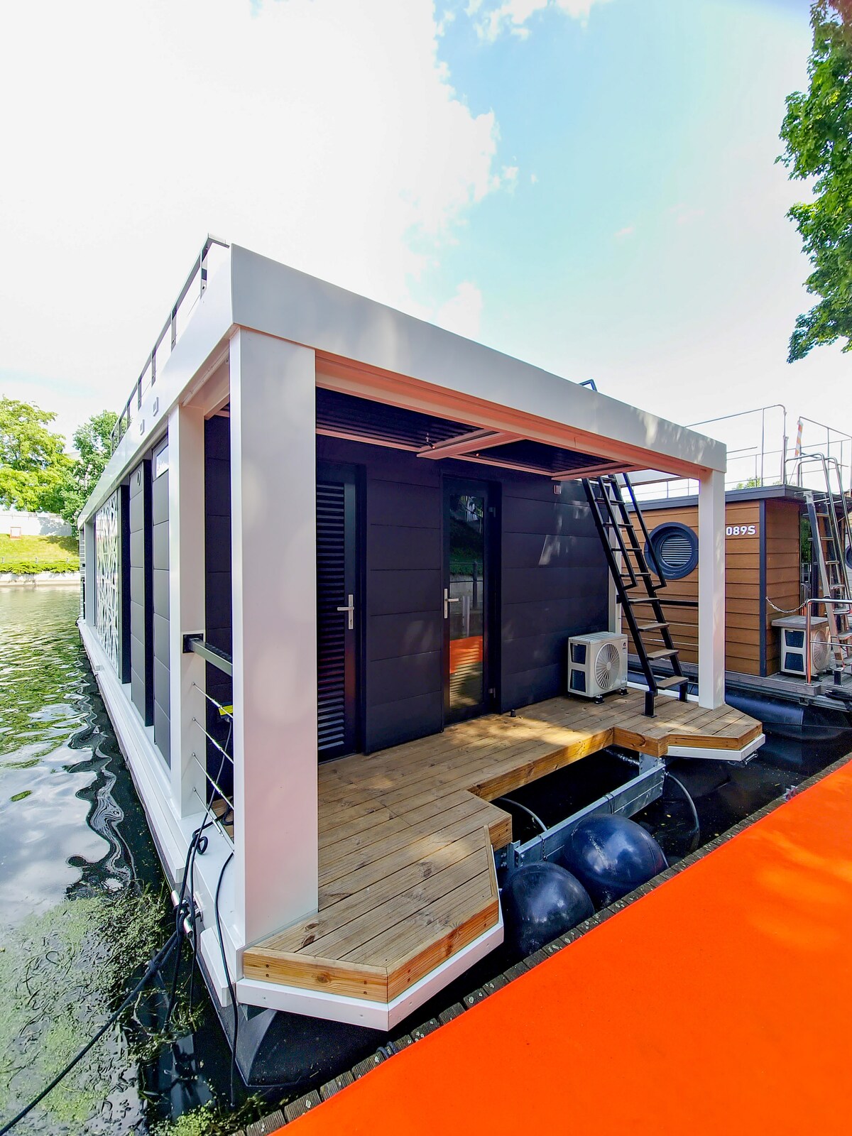 The houseboat exterior features a modern design with a wooden deck leading to the entrance. Large windows allow for natural light, while a ladder provides access to the upper deck. The surrounding water reflects the vibrant blue sky and greenery.