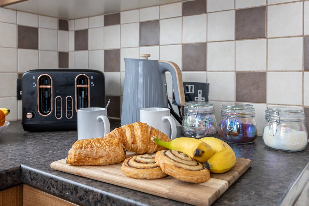 A kitchen countertop displays a wooden board with a variety of breakfast items, including pastries and bananas. Two mugs sit beside a kettle and a toaster, while jars filled with colorful contents are arranged in the background.