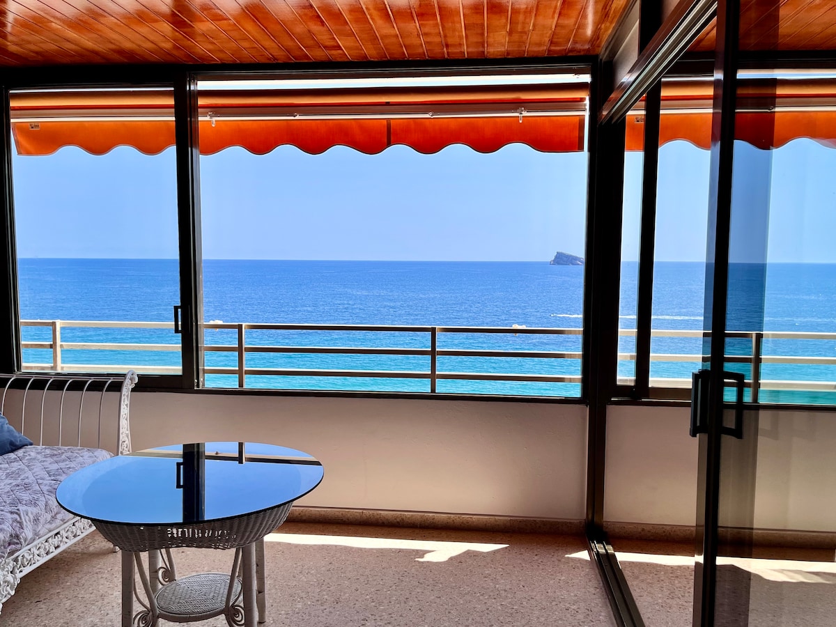 A sunlit terrace features expansive glass windows providing unobstructed views of the coastline and sea. A round glass table is positioned in the center, surrounded by a white metal framework bench. Bright blue waters meet the sky in the distance, enhancing the sense of openness.