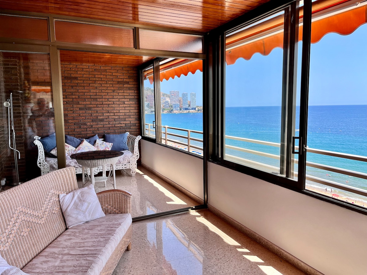 A bright living space features large glass windows that offer panoramic views of the beach and ocean. Comfortable seating is arranged around a round table, with soft textiles adding a relaxed ambiance. Natural light fills the area, enhancing the spacious feel.