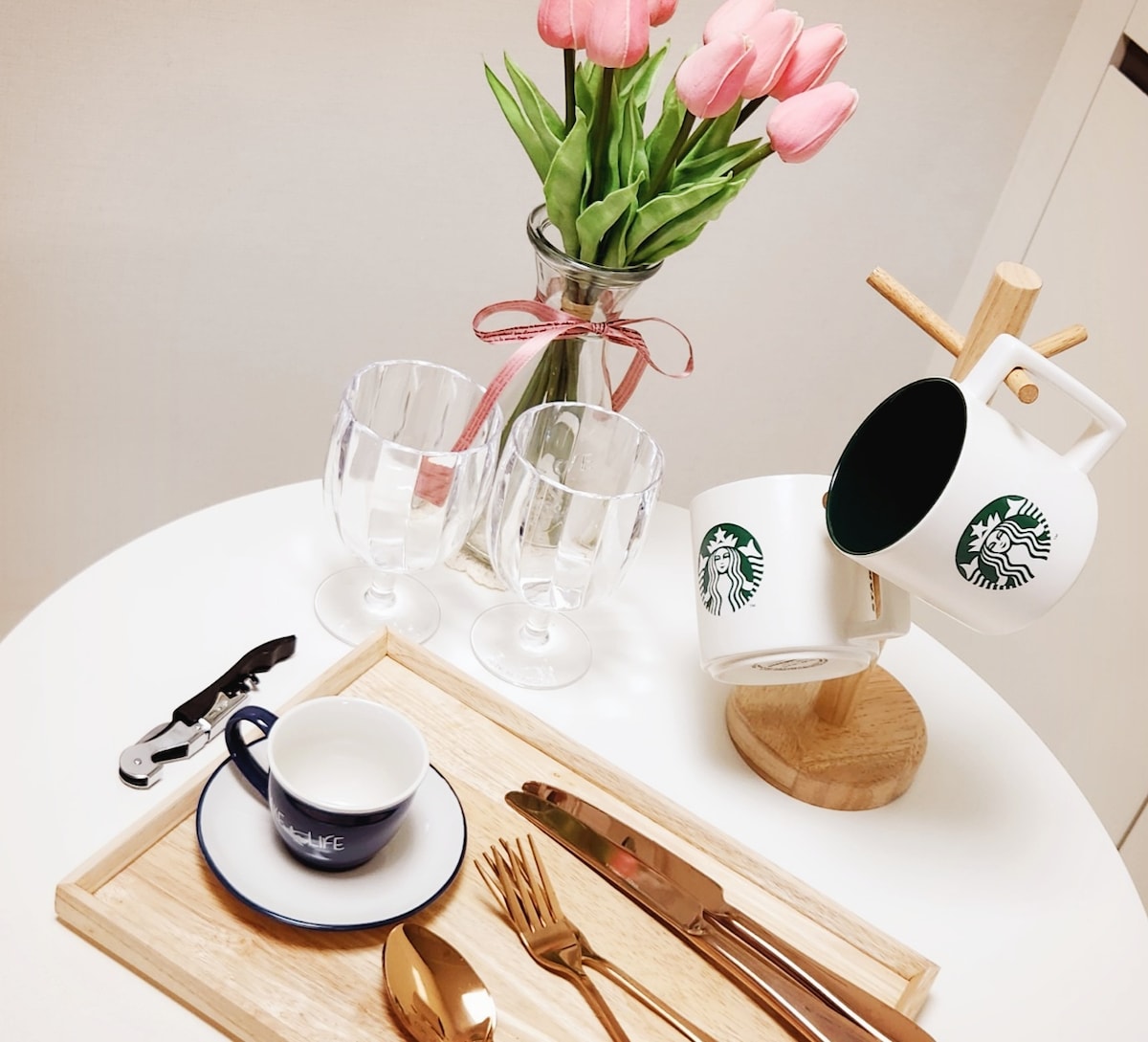 A round table is set with various tableware, including a wooden tray, mugs, and glassware. A vase with fresh pink tulips adds a touch of color, while silver cutlery and a coffee tool are arranged neatly, creating an inviting dining experience.
