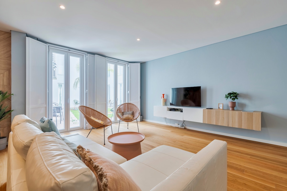 A bright living area is presented, featuring a light-colored sofa facing a modern, wall-mounted TV. Two stylish chairs are positioned near large windows, allowing natural light to fill the space. A small circular table sits in the center, enhancing the open and airy feel.