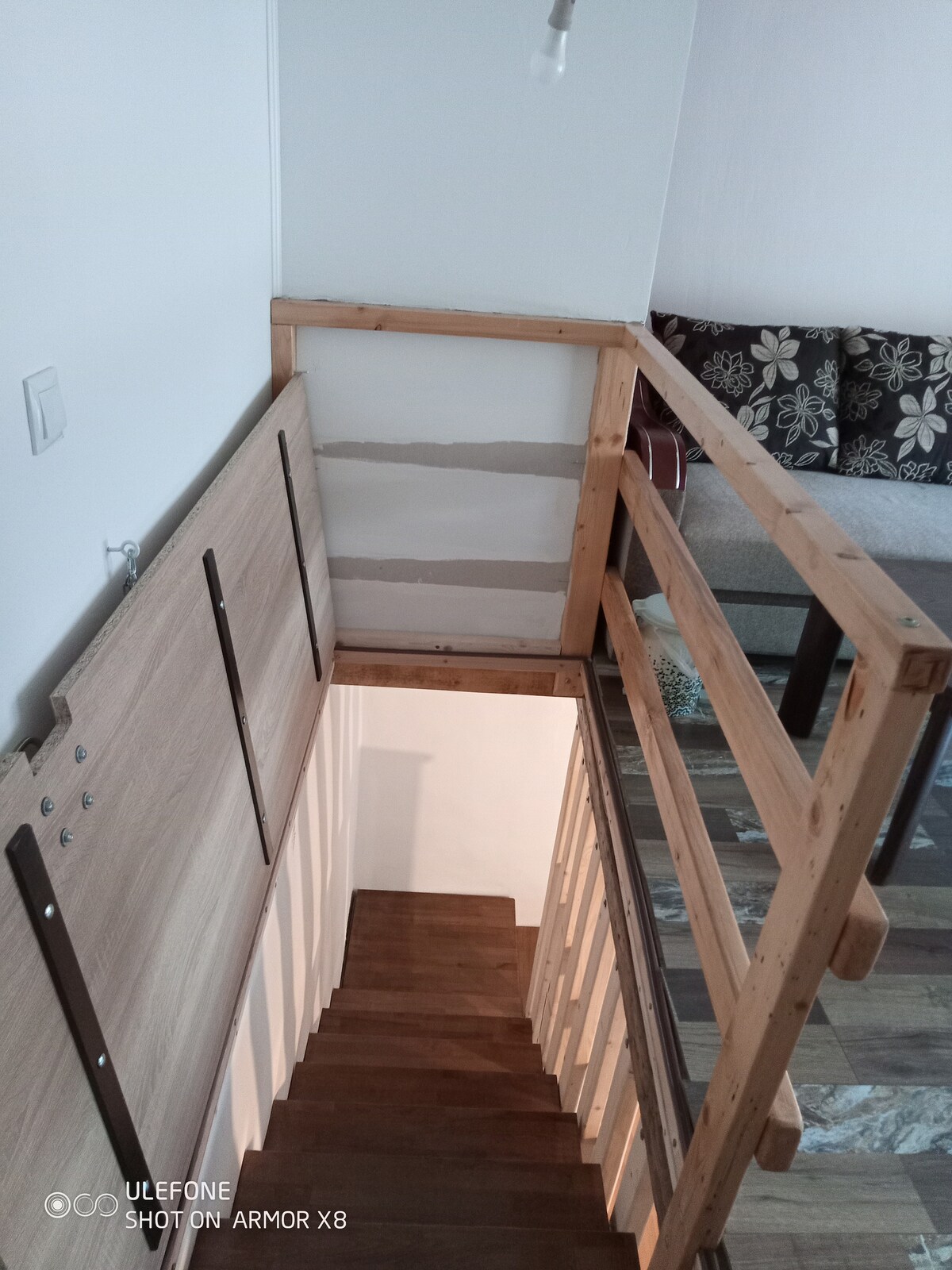 A wooden staircase with a light-colored railing leads downwards. Natural wood steps are visible, complemented by a well-lit space below. A cozy seating area with decorative cushions is seen in the background, providing hints of comfort and style.