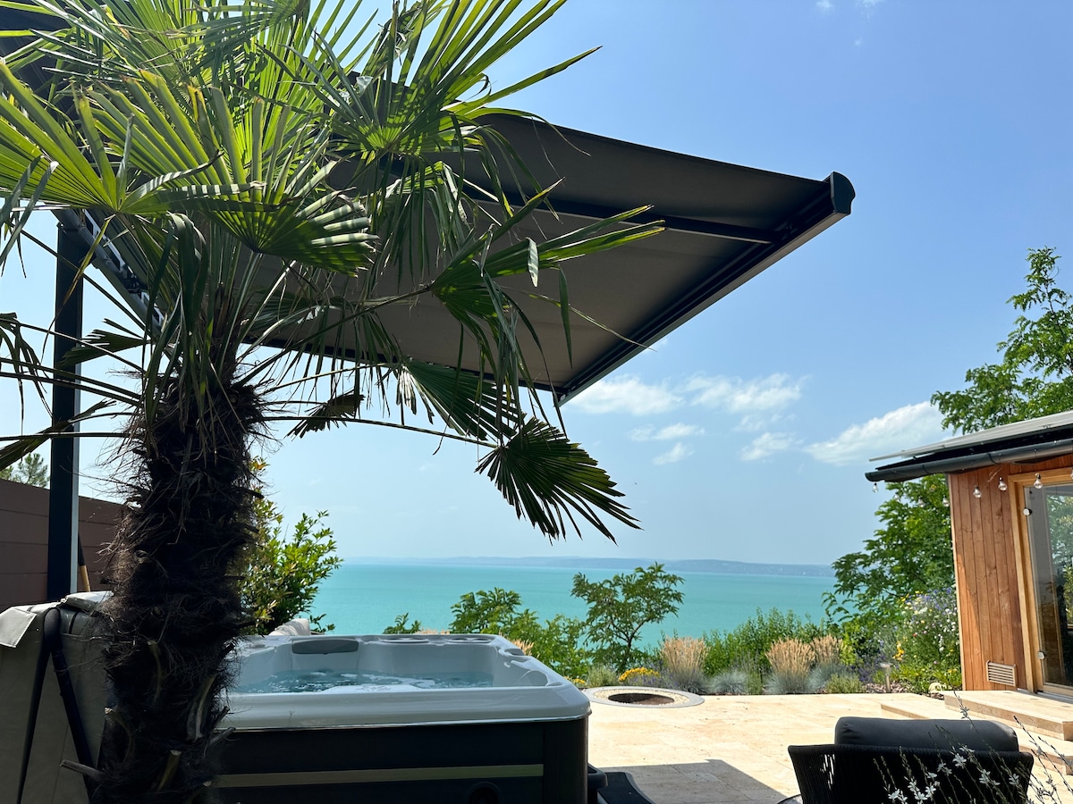 A view of the serene Balaton landscape is framed by a palm tree and a retractable awning. The outdoor space features a jacuzzi, with the lake visible in the background against a clear blue sky. Lush greenery surrounds the area, enhancing the tranquil setting.
