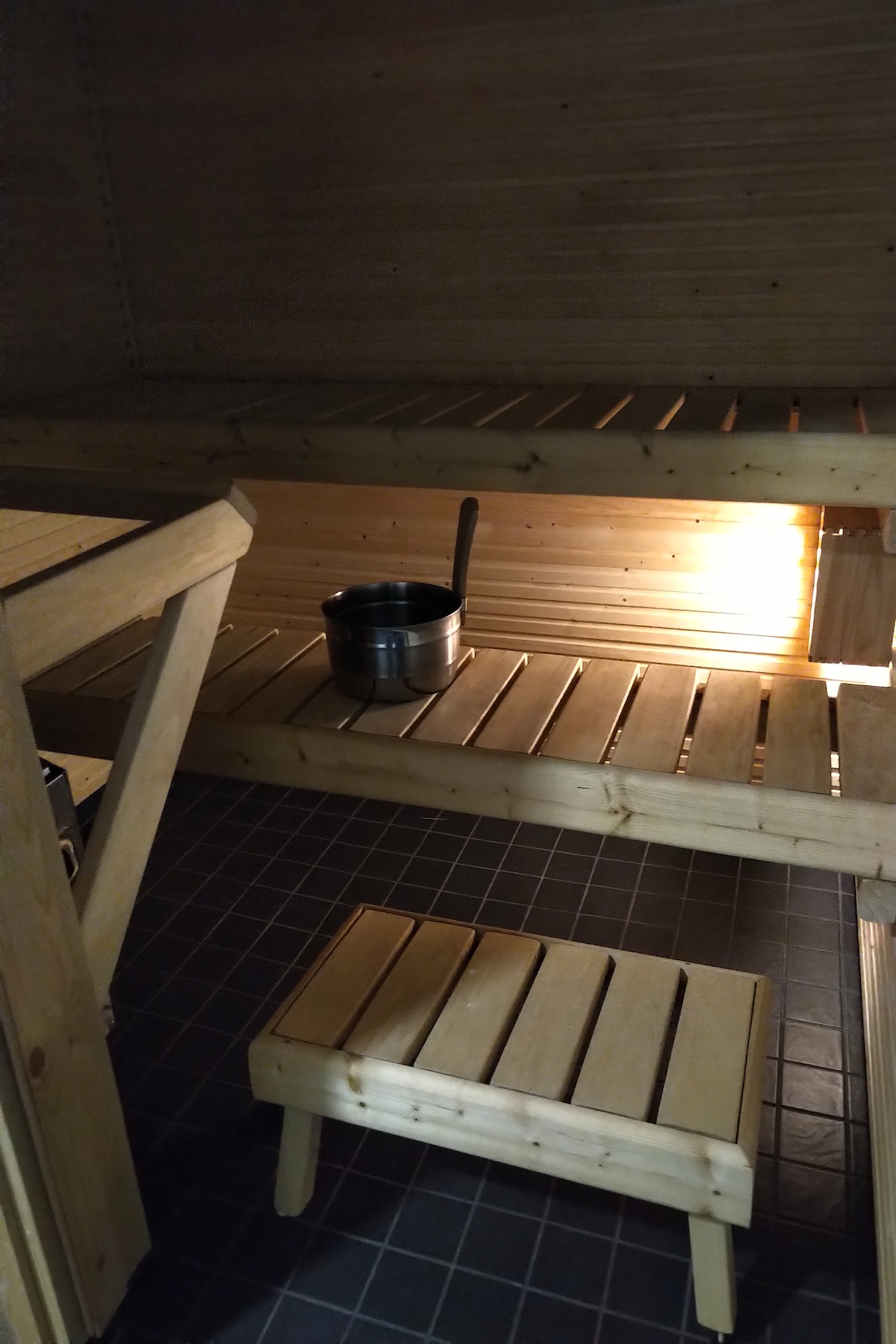 A wooden sauna features multiple bench levels, with a small stool positioned on the floor. A stainless steel sauna bucket is located on one of the benches, with warm light illuminating the wooden interior.