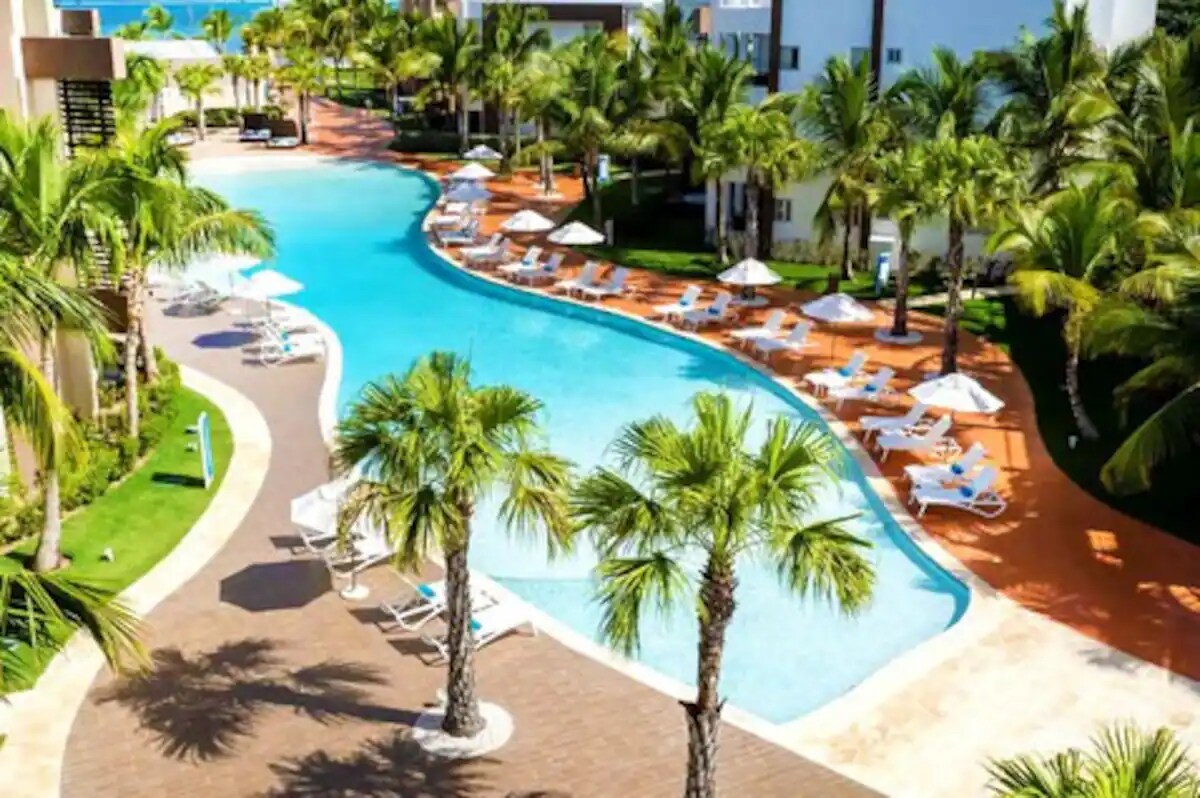 A curved swimming pool is lined with sun loungers and umbrellas, surrounded by lush palm trees. The vibrant pathway leads to beautifully landscaped areas, creating an inviting space for relaxation and leisure, with glimpses of the ocean in the distance.