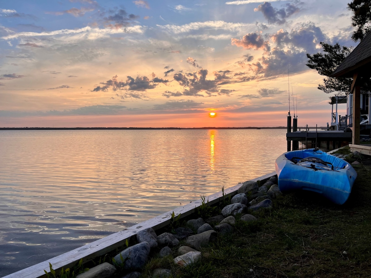 A clear evening view of the water showcases a vibrant sunset reflecting on the surface. A blue kayak rests on the grass beside a stone-lined edge, with gentle waves lapping against the shore. Soft clouds accentuate the colorful sky, creating a serene atmosphere.