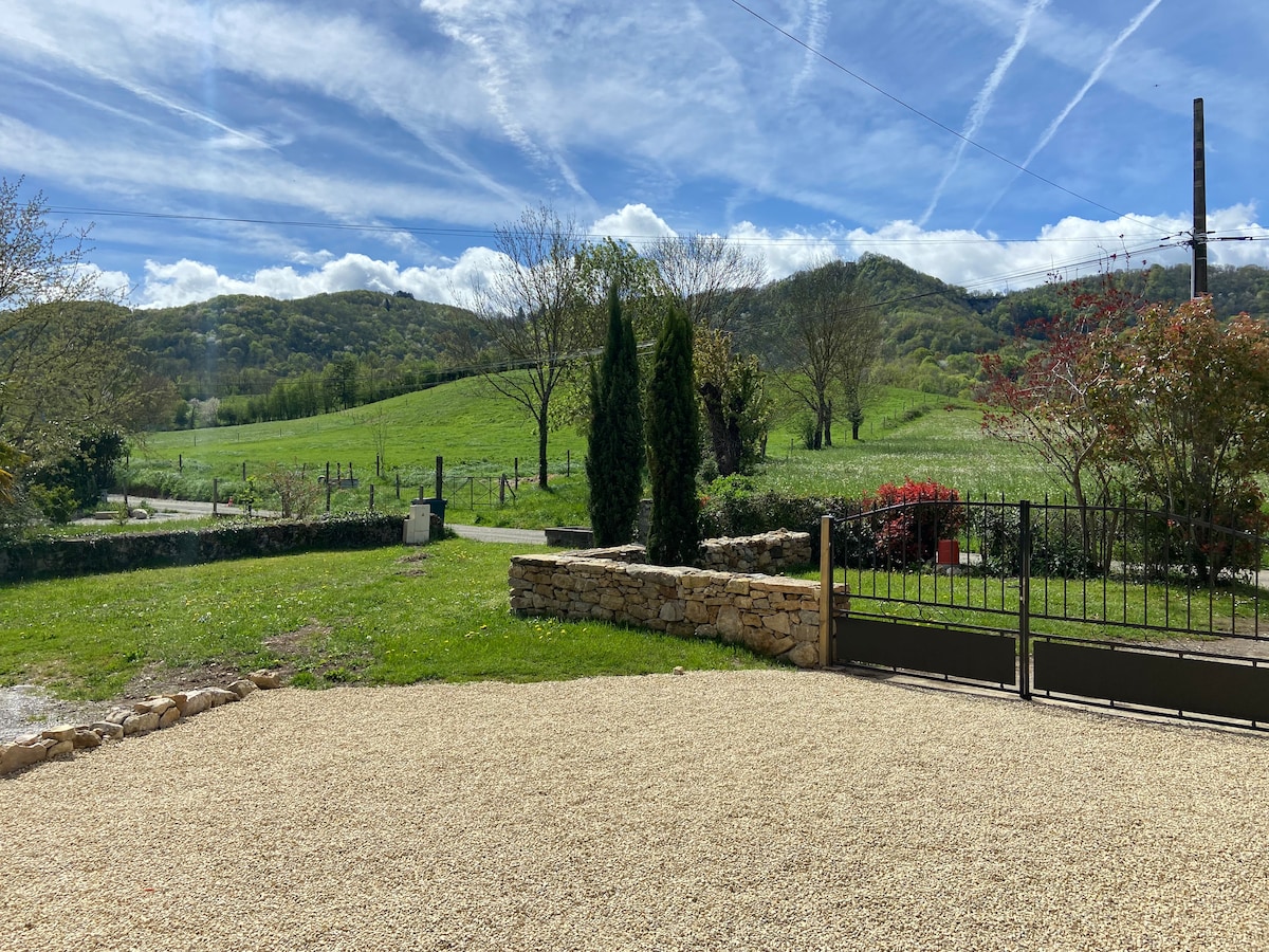 A scenic view of a green landscape is captured, featuring rolling hills and a clear blue sky with clouds. A stone wall and gate frame the foreground, while trees and shrubs add to the natural surroundings.