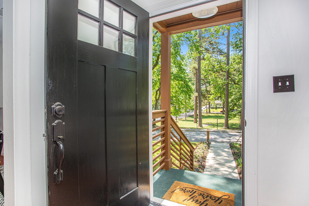The entrance showcases a stylish black door with a grid window, leading to a welcoming pathway surrounded by greenery. Sunlight filters through the trees, highlighting the inviting outdoor space. A doormat with a friendly message adds a personal touch to the entryway.