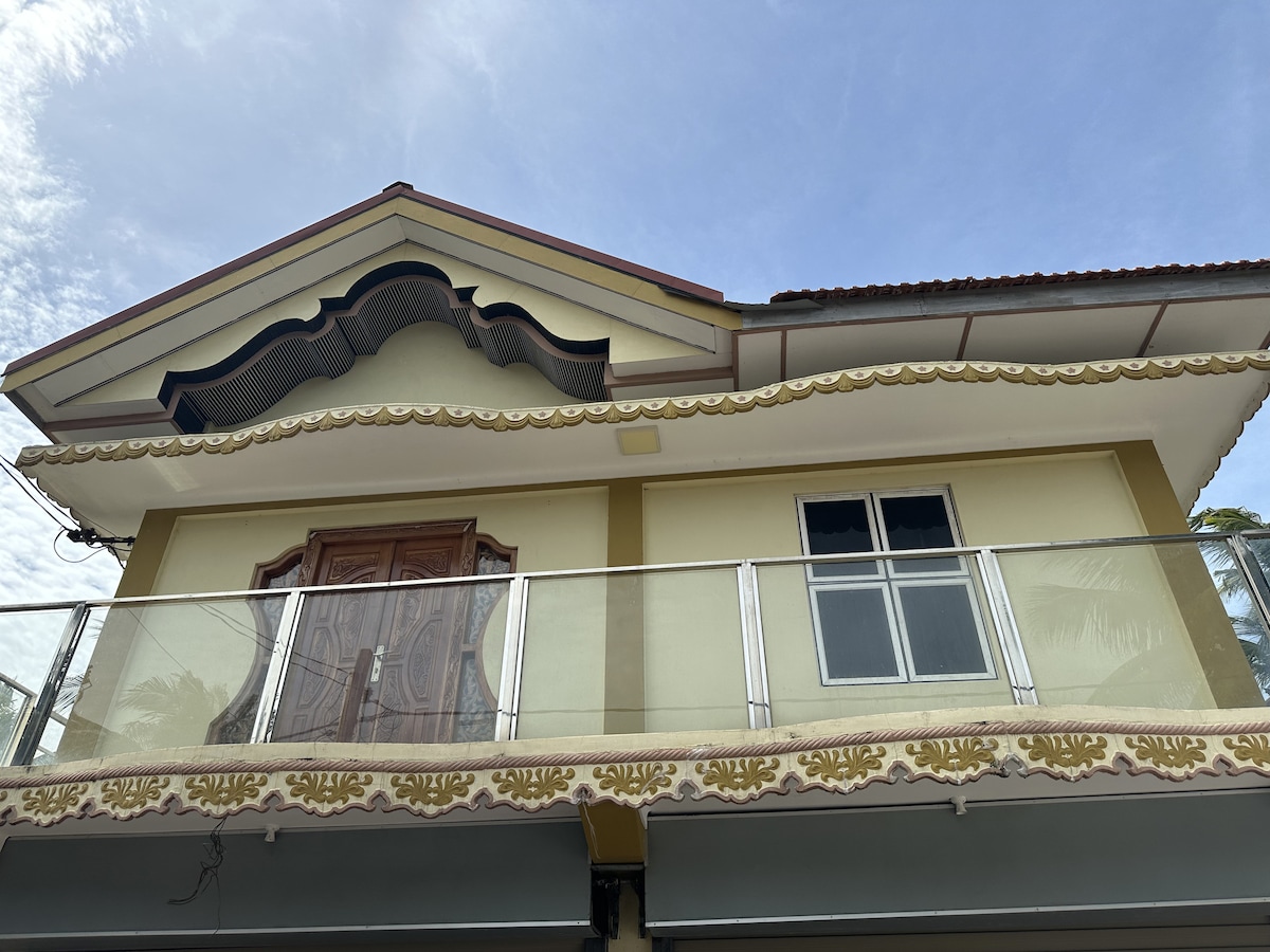 The exterior of the house showcases traditional architectural details, including ornate patterns along the eaves and a spacious balcony. Large windows allow for natural light, and the wooden front door features intricate carvings, adding character to the inviting facade.
