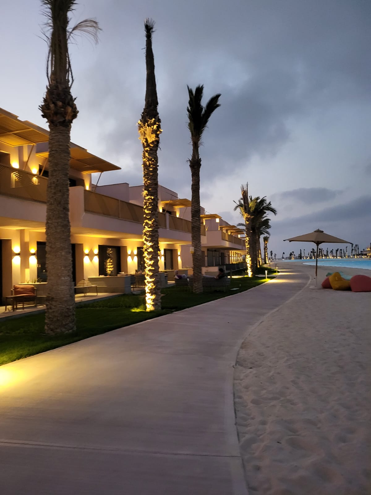 A serene beachfront pathway is lined with palm trees, their trunks illuminated by warm lighting. The walkway leads to beachfront villas, with soft light spilling from their façades, enhancing the evening ambiance. Comfortable seating areas are visible along the path, inviting relaxation.