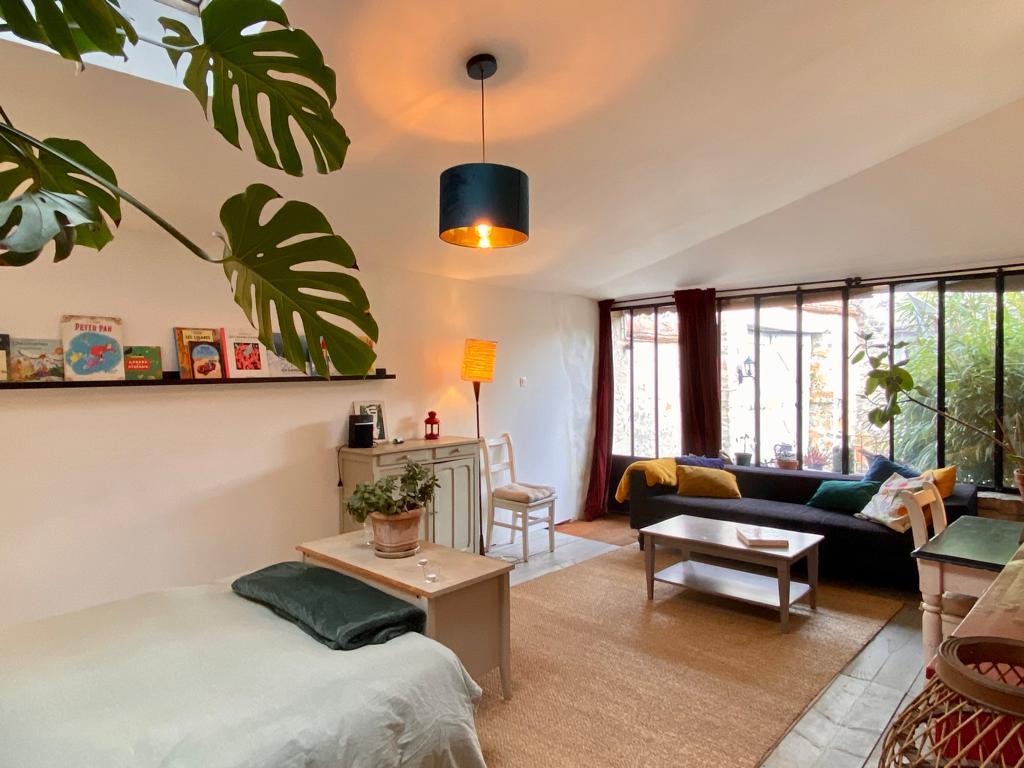 A contemporary living space features a cozy sleeping area with a soft bed and simple bedding. Natural light streams in through large windows, illuminating a green sofa and a small dining area accompanied by minimalistic decor. Potted plants add a touch of nature to the room.