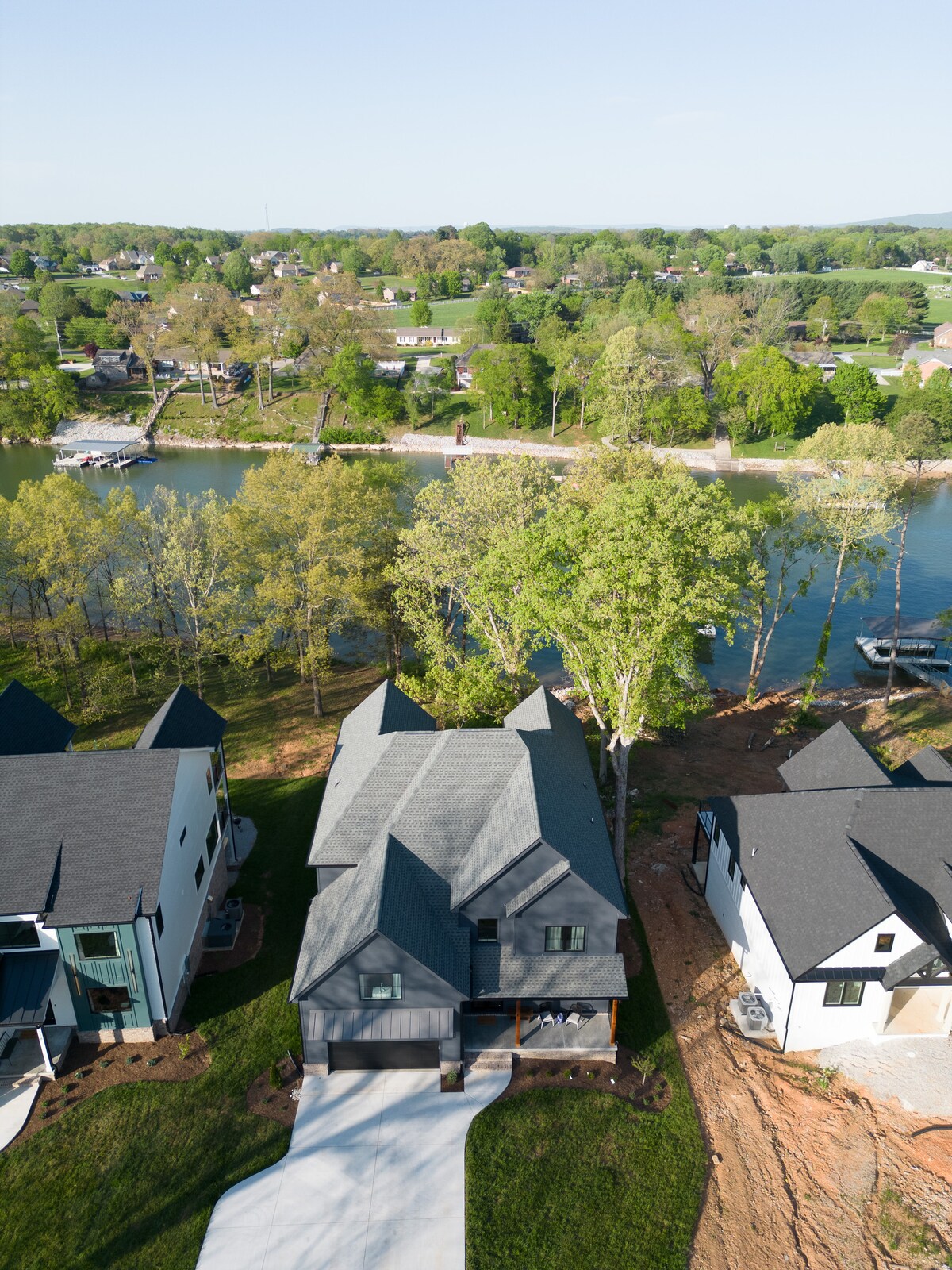An aerial view captures Kayak Kove, a dark grey house with a paved driveway, surrounded by lush greenery. The property is adjacent to a serene lake, with boats visible along the shoreline and trees framing the scene. Nearby homes with dark roofs complete the picturesque neighborhood.