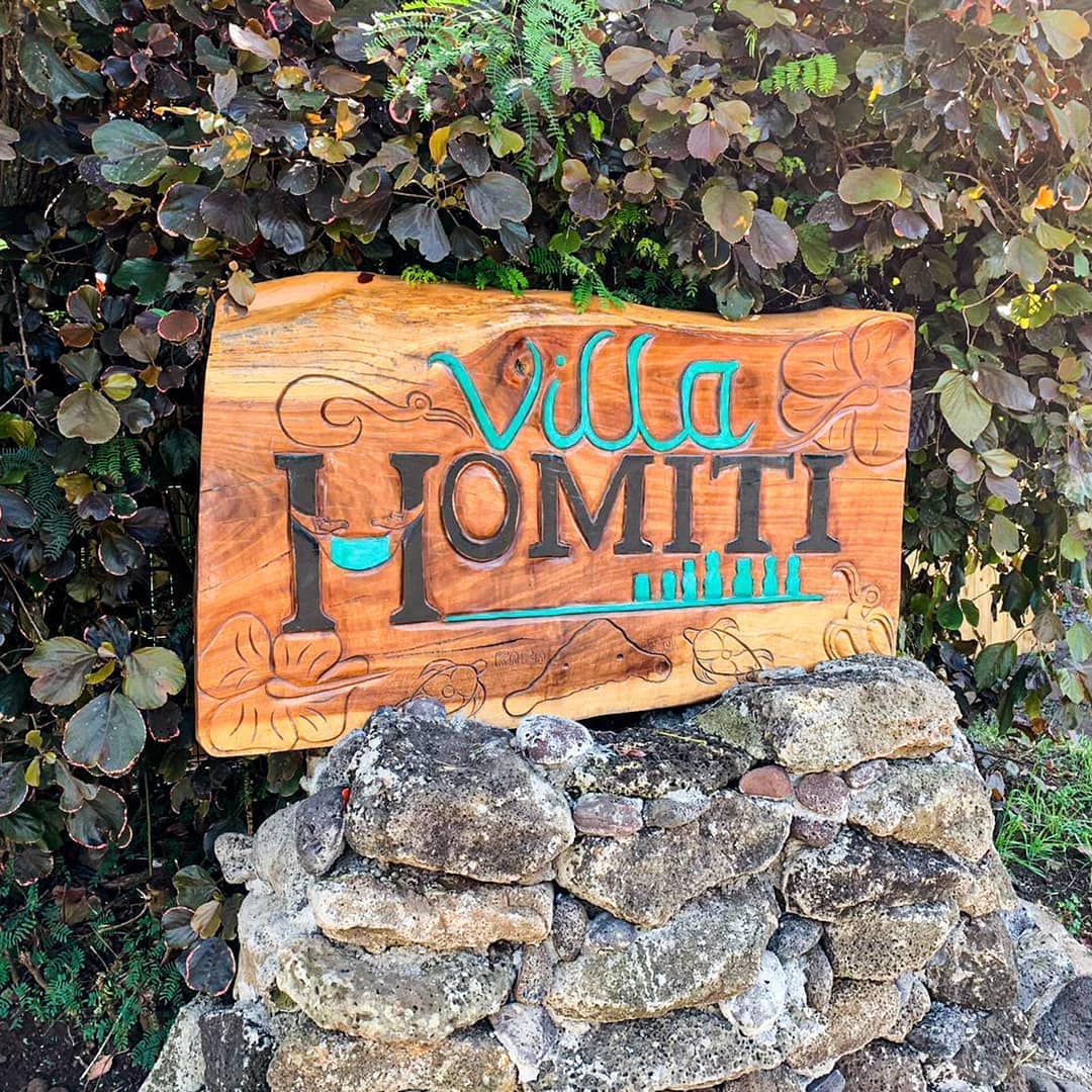 A handcrafted wooden sign displaying the name 'Villa Homiti' is situated on a natural stone base. The backdrop features lush green foliage, contributing to a welcoming entrance to the property.