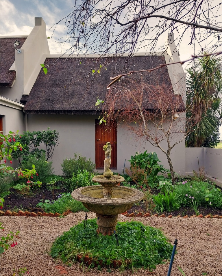 A quaint garden scene features a stone fountain at its center, surrounded by lush greenery and colorful flowers. The thatched-roof building is visible in the background, with wooden doors inviting a sense of warmth and tranquility. A tree branches overhead, adding natural charm.