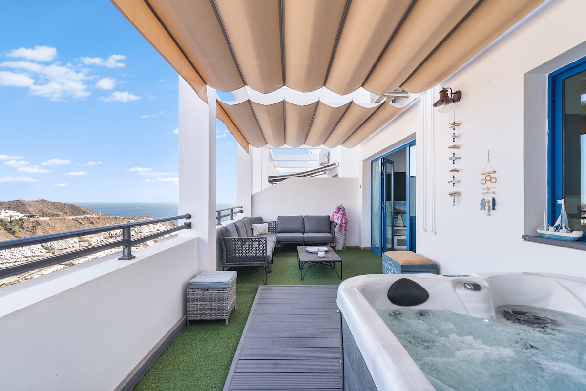 A private terrace is presented, featuring a jacuzzi and a comfortable seating area with a sofa and chair set. The space is partially shaded by an awning, and scenic ocean views can be seen in the background, enhancing the space's tranquility.