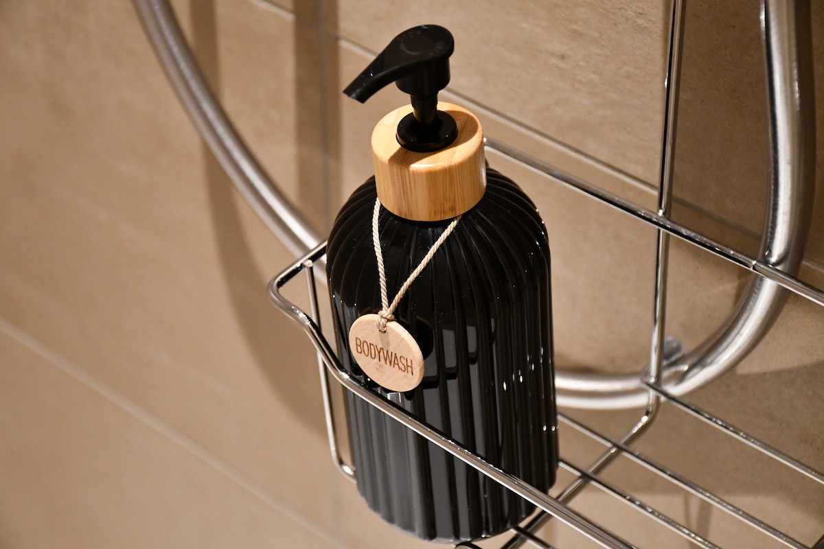 A black bottle with a wooden pump is positioned on a metal shower shelf. A tag labeled 'BODYWASH' hangs from the bottle, which is set against a neutral tiled wall.