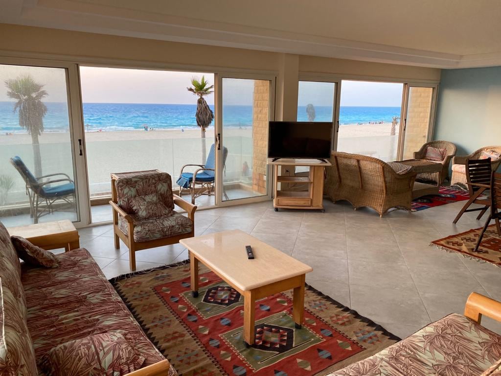 A spacious living area is shown with large glass doors, providing a view of the beach. Comfortable seating is arranged with tropical-patterned cushions, while a flat-screen TV sits atop a wooden stand. A dining table and chairs complement the room's decor.