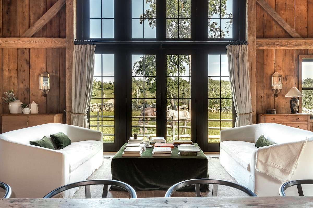 The living area features two light-colored sofas positioned around a low coffee table stacked with books. Large windows provide a view of the green landscape outside, framed by wooden beams and soft, flowing curtains.