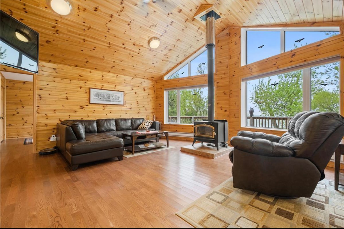 A spacious living area features warm wooden walls and large windows, allowing natural light to fill the space. A comfortable dark leather sectional sofa is arranged around a central area, with a cozy chair positioned nearby. A wood stove adds to the cabin's inviting atmosphere.