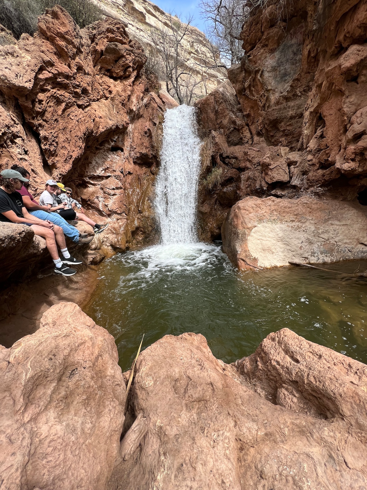 A flowing waterfall cascades over rocky terrain, creating a serene pool at the base. Surrounding the water are rugged cliffs and boulders, with sparse vegetation partially visible. Two individuals are seated on the rocks, enjoying the natural setting.